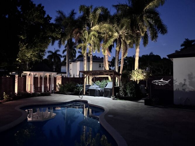 A backyard at dusk featuring a swimming pool, palm trees, patio furniture under a pergola, and a lit wall with a fish decoration.
