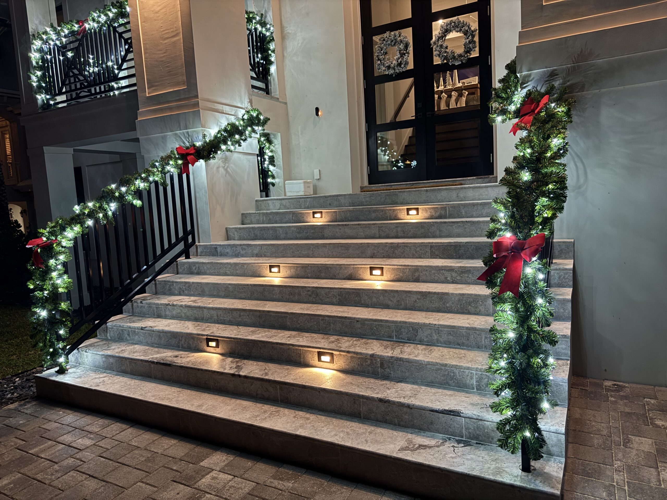 Front steps of a house decorated with lit garlands and red bows for the holidays; wreaths hang on the glass front doors, and step lights illuminate the stairs.