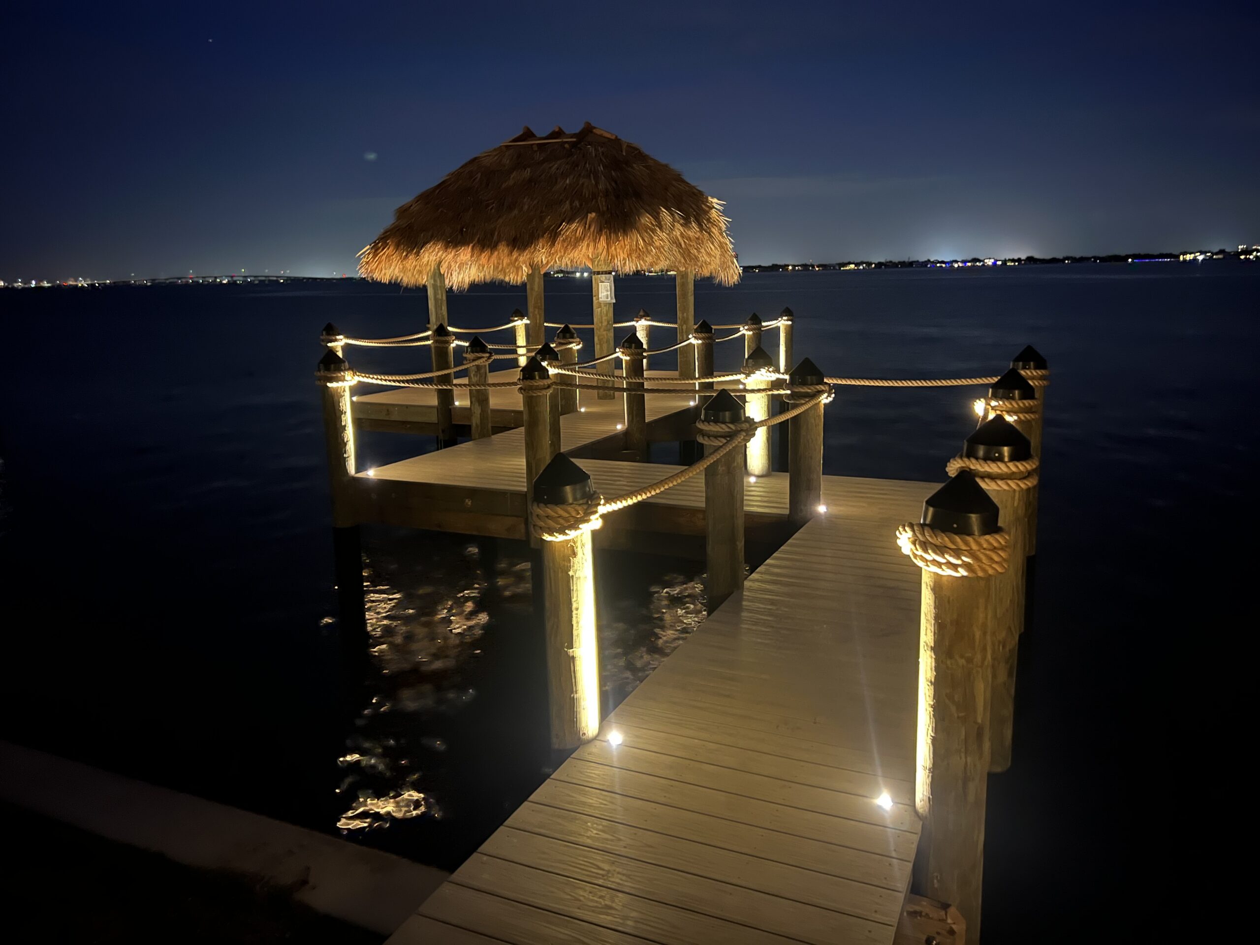A wooden pier with rope railings and a thatched-roof gazebo extends over calm water at night, illuminated by soft lights.