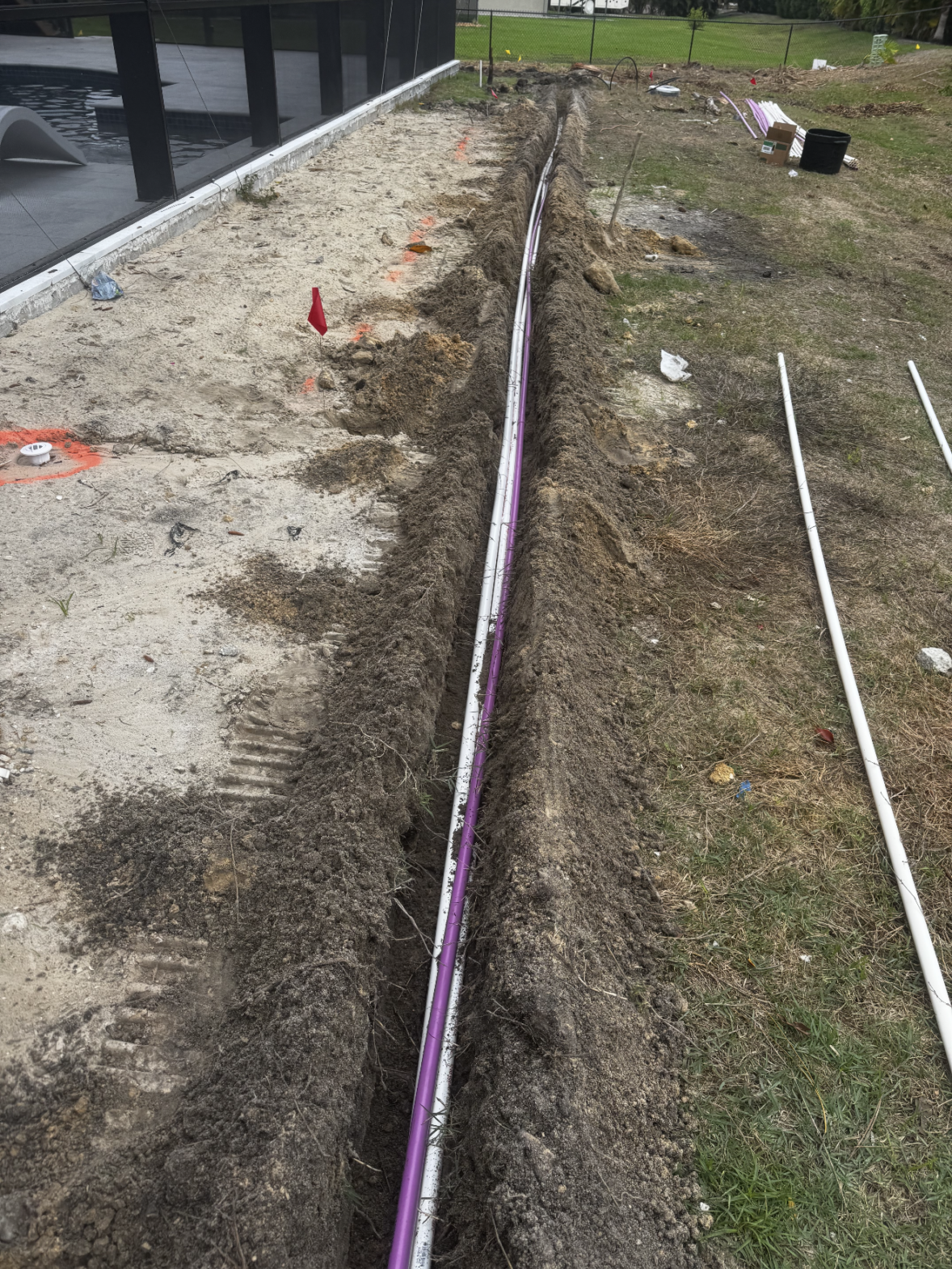 A purple conduit is laid in a narrow trench next to a building under construction, with dirt and construction materials visible nearby—an essential step often overseen by a landscape lighting designer to ensure seamless integration of exterior lighting systems.