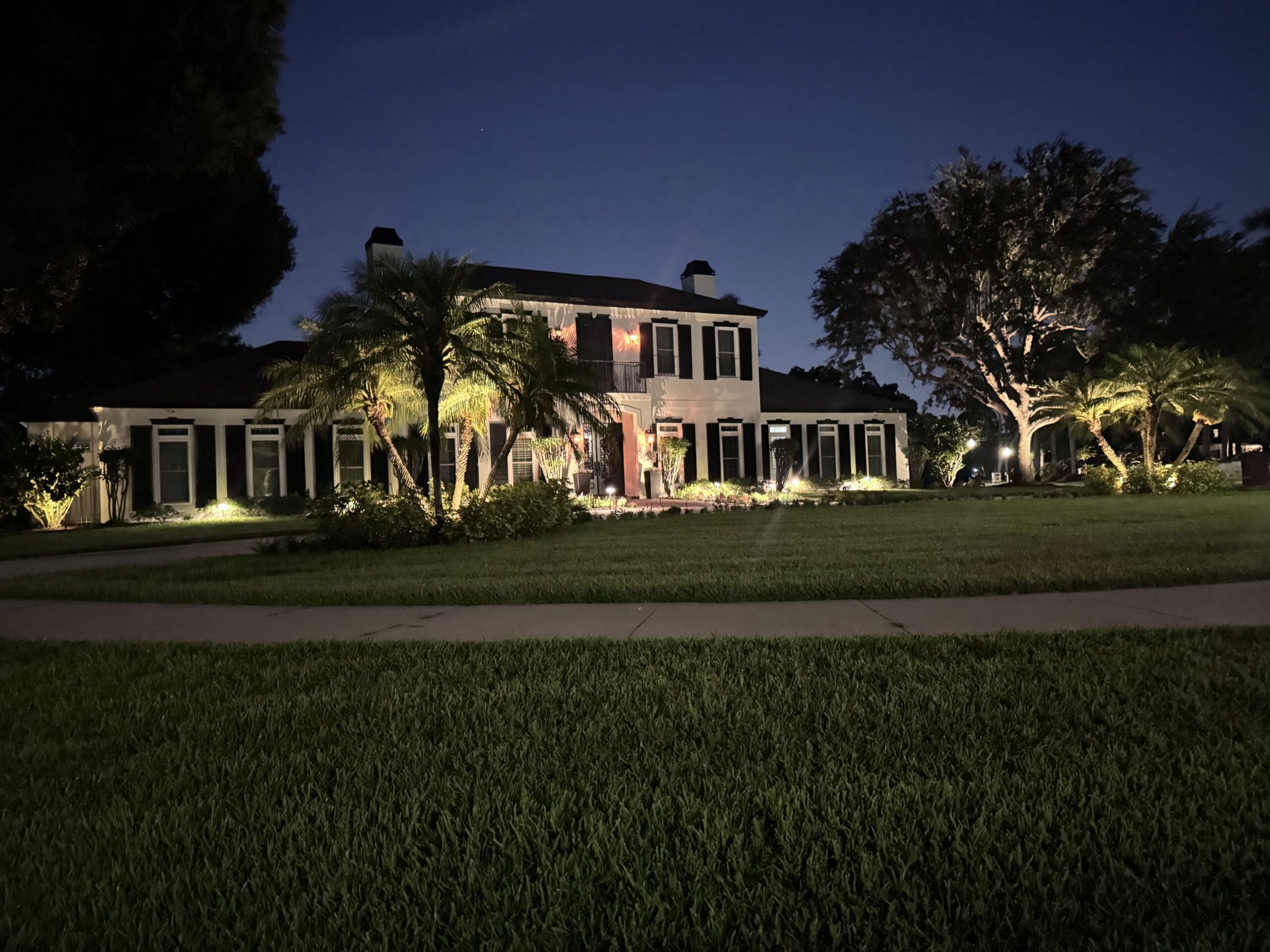 Two-story white house with black shutters and palm trees in front, illuminated by outdoor lights at night, with a large lawn and sidewalk in the foreground.