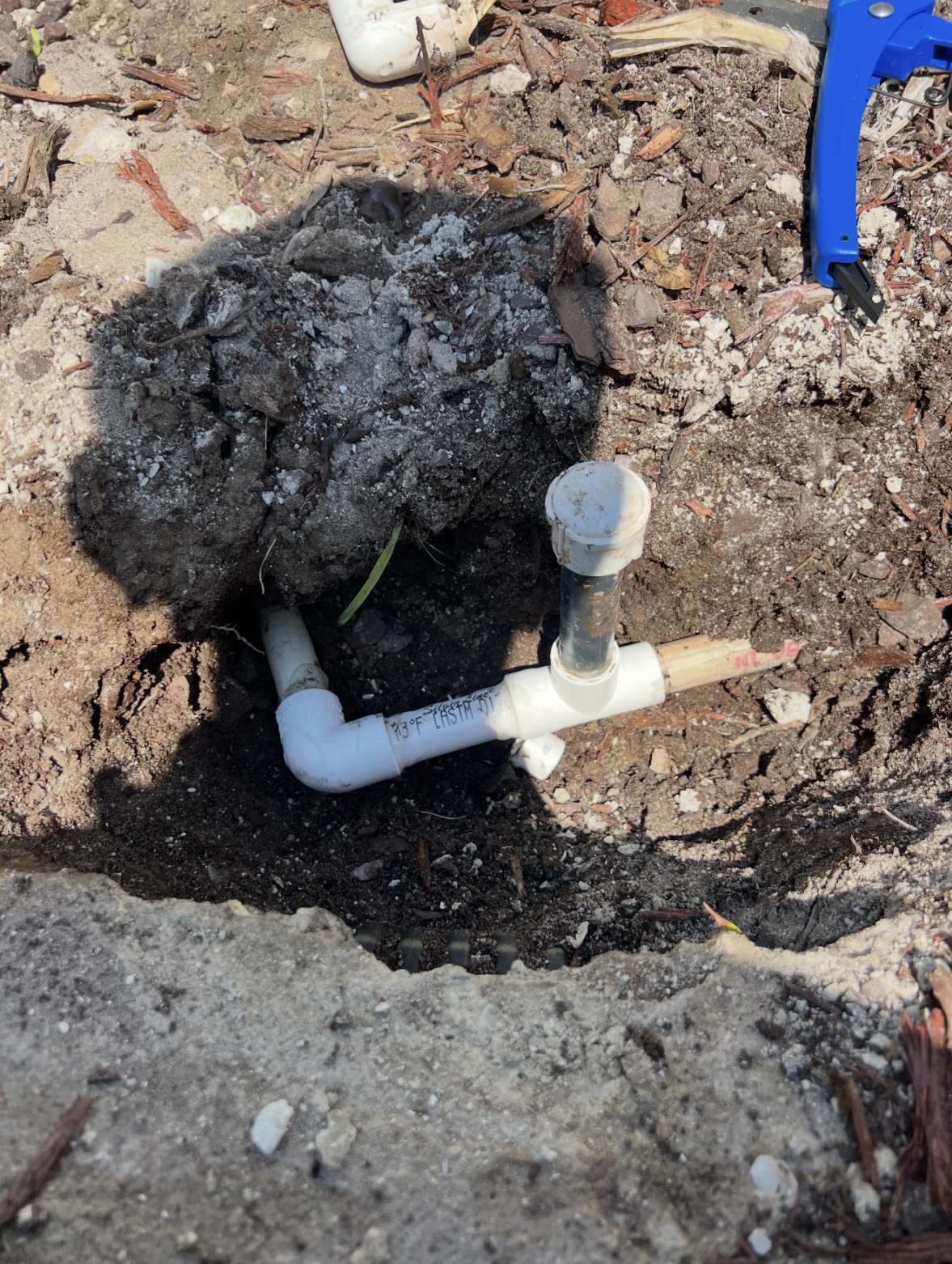 Exposed PVC irrigation pipe with an attached sprinkler head is partially buried in soil and surrounded by dirt, debris, and a few tools in the background—details that often draw the eye of a landscape lighting designer when planning seamless outdoor illumination.