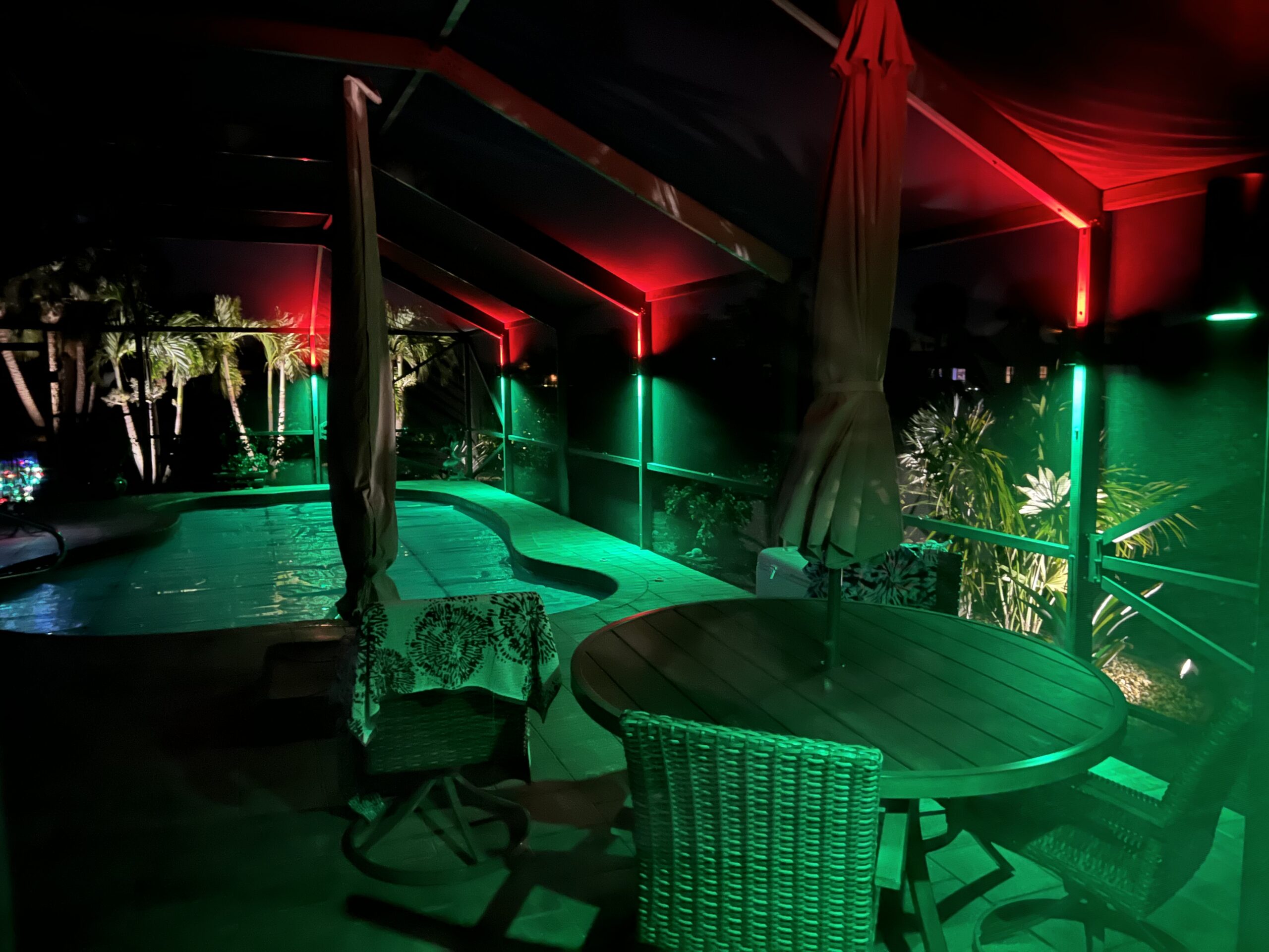 Covered patio at night with a round table, chairs, and closed umbrellas beside a swimming pool, illuminated by red and green accent lights.