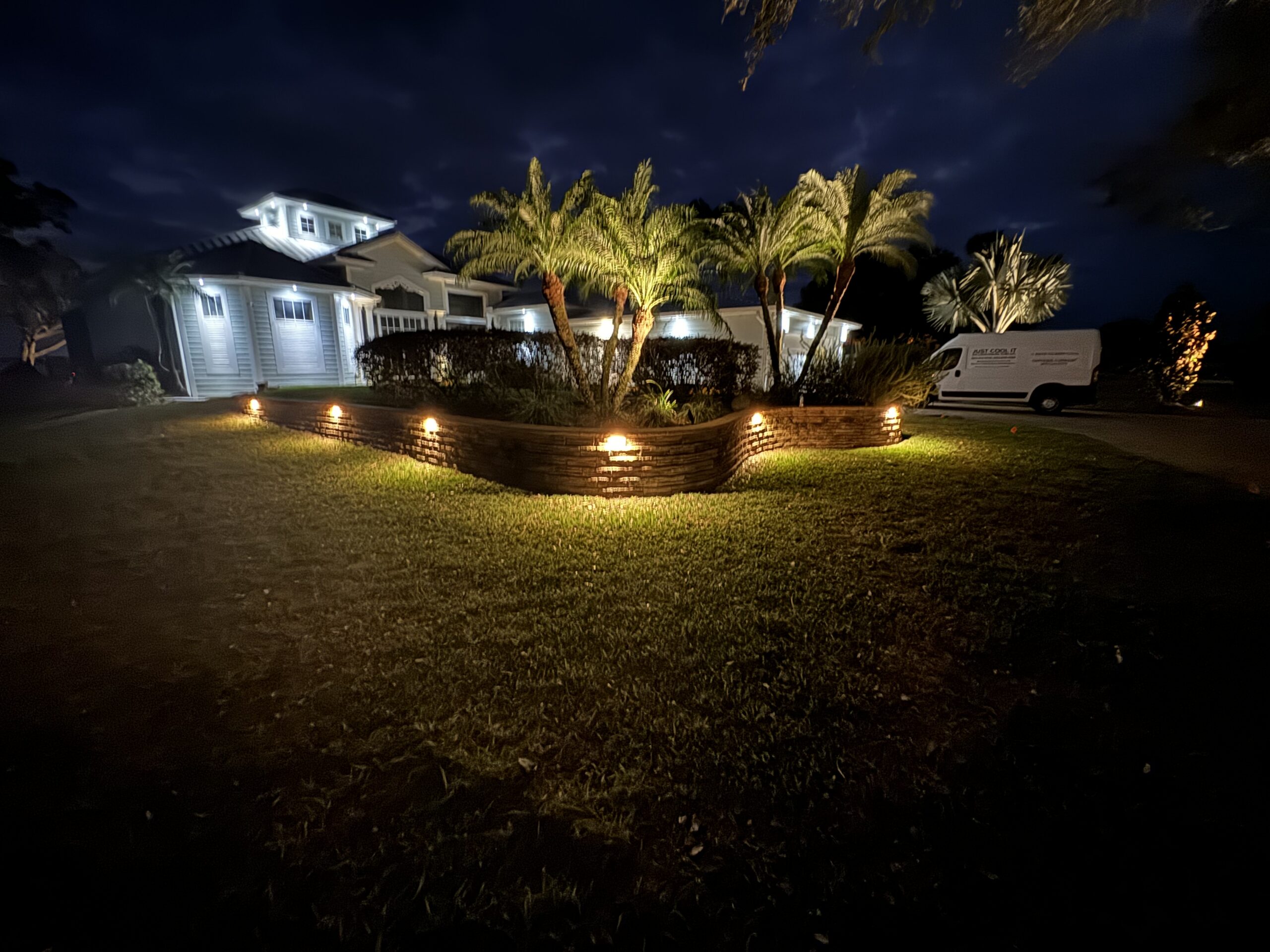 A house at night with outdoor lights illuminating palm trees and a stone retaining wall; a white van is parked in the driveway.