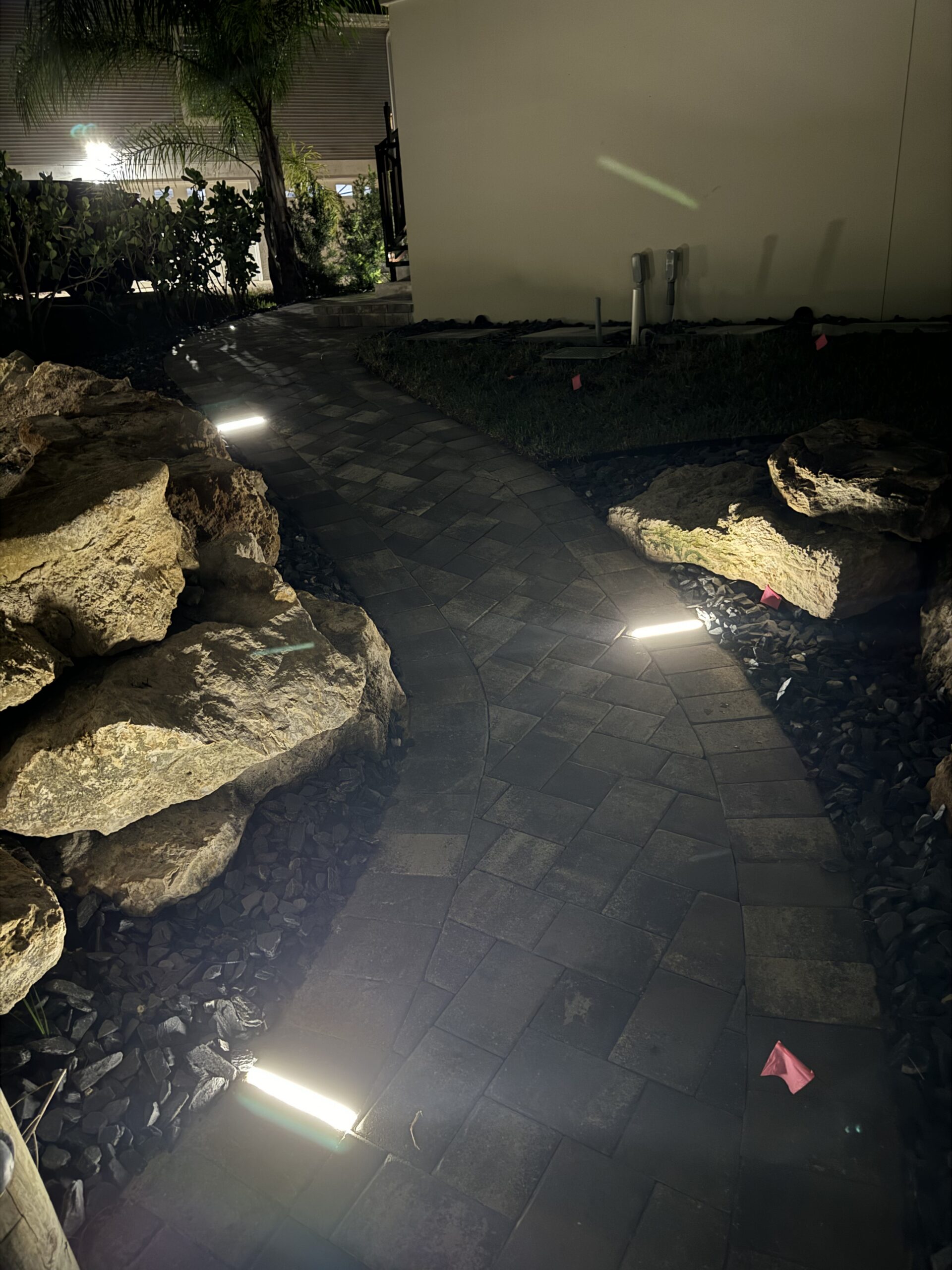 A stone walkway curves between rocks at night, illuminated by several rectangular ground lights, with pink flags marking spots on the ground.