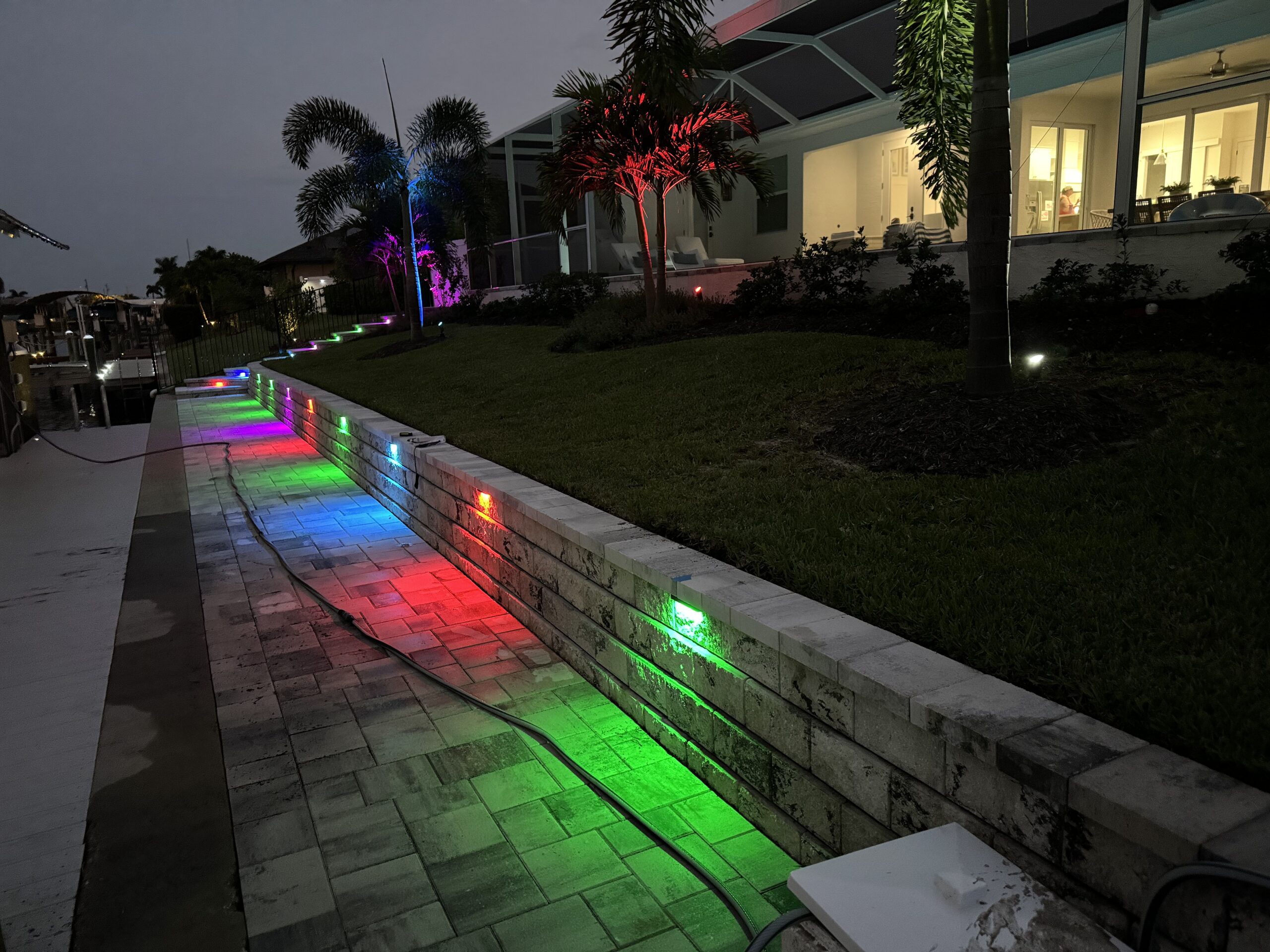Colorful LED lights illuminate a backyard retaining wall and walkway at dusk, with a modern house and palm trees in the background.