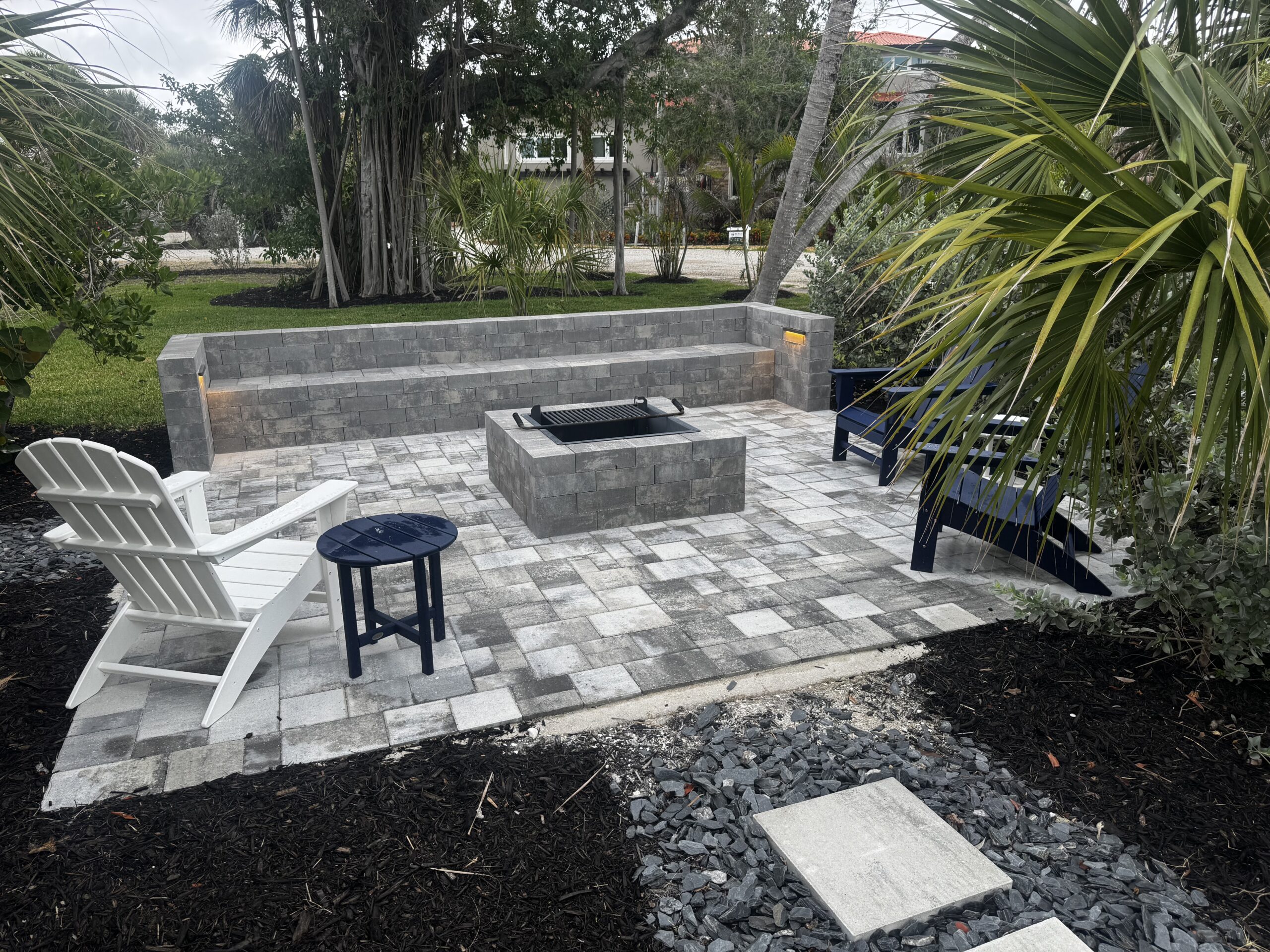 Outdoor patio with gray stone pavers, a built-in bench, a central fire pit, and four chairs surrounded by greenery and palm trees, thoughtfully illuminated by a landscape lighting designer to create an inviting ambiance after sunset.