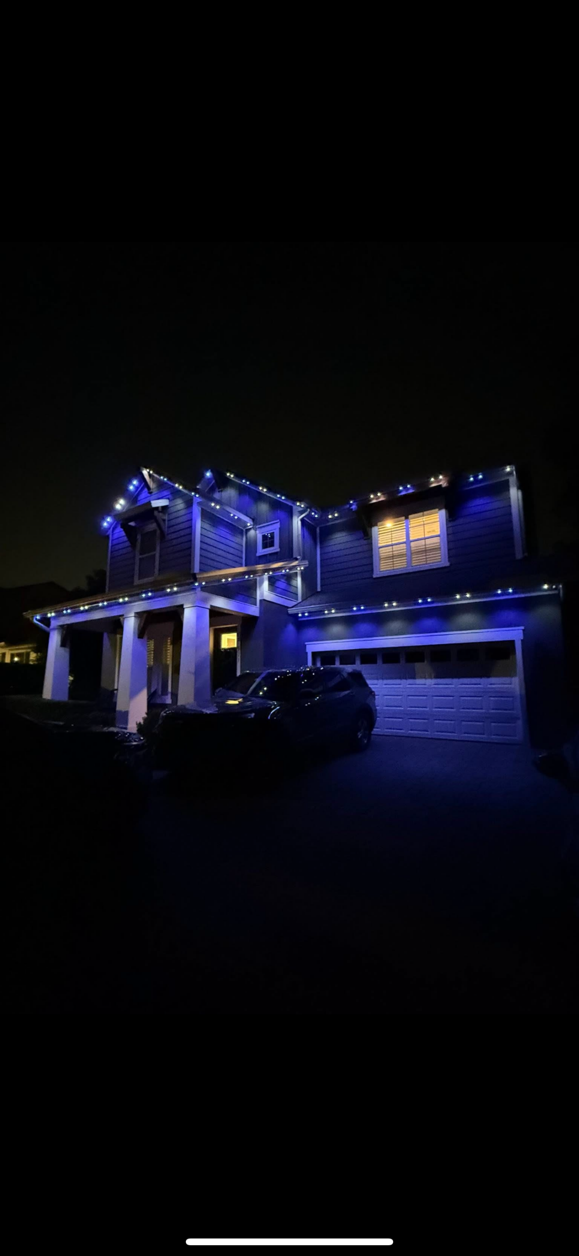 A two-story house at night with blue and white string lights outlining the roof and garage, thoughtfully arranged to highlight the home's features like a landscape lighting designer would. A dark-colored car is parked in the driveway.