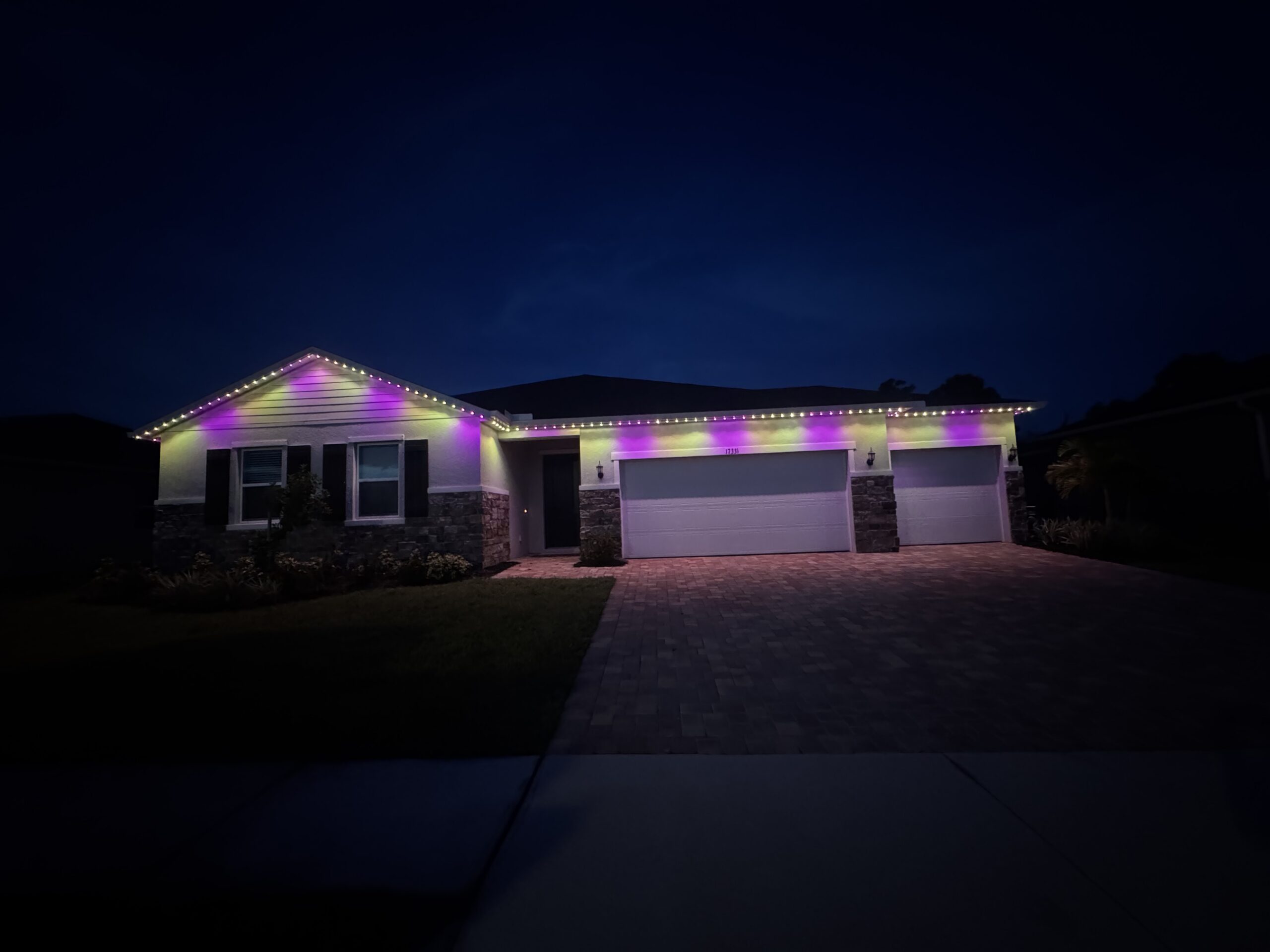 Single-story house at night with purple, yellow, and white permanent lighting along the roofline; three-car garage and brick driveway are visible.