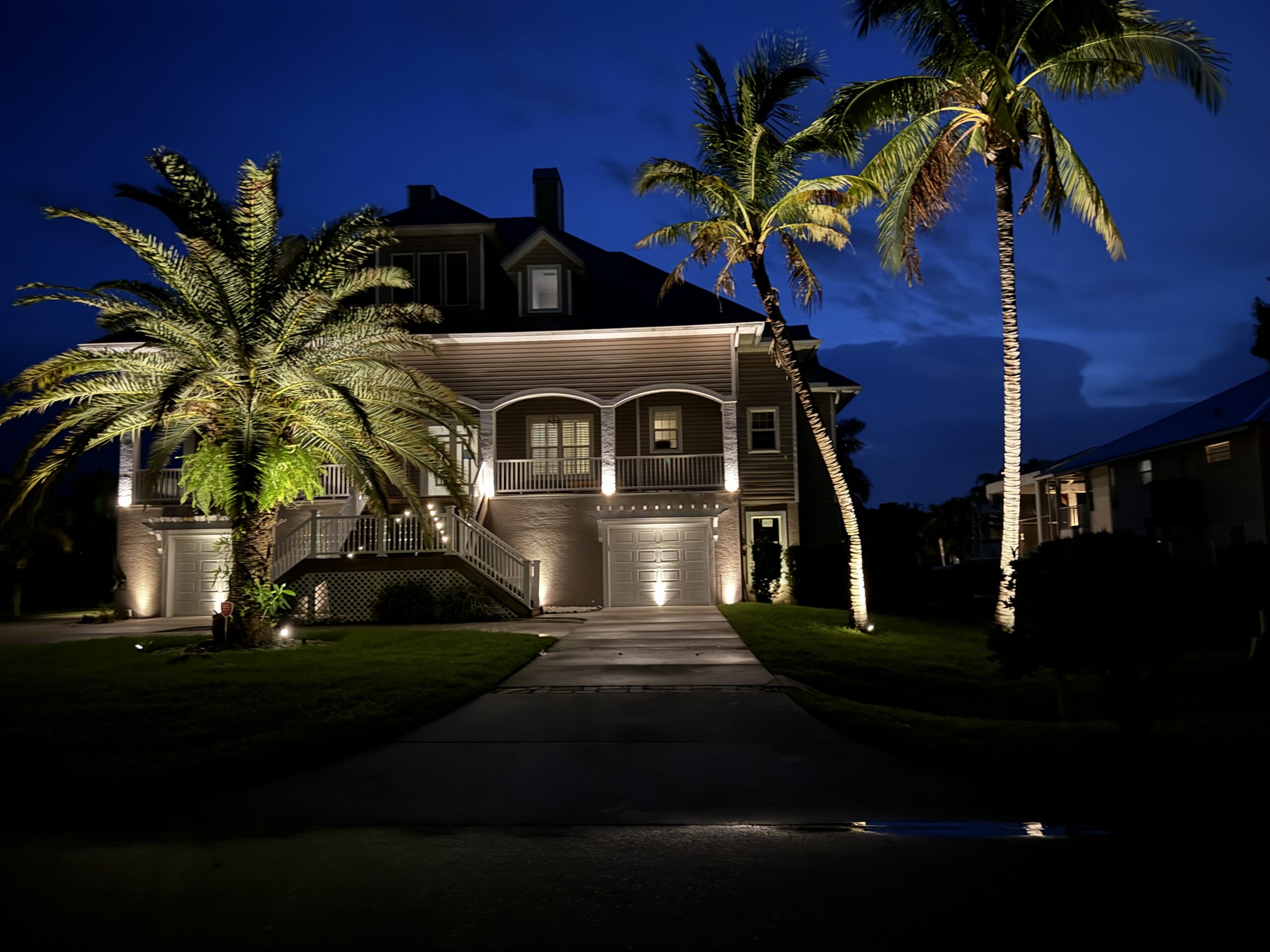 A large, two-story house with exterior lights is surrounded by palm trees at night, with a driveway leading to the garage.