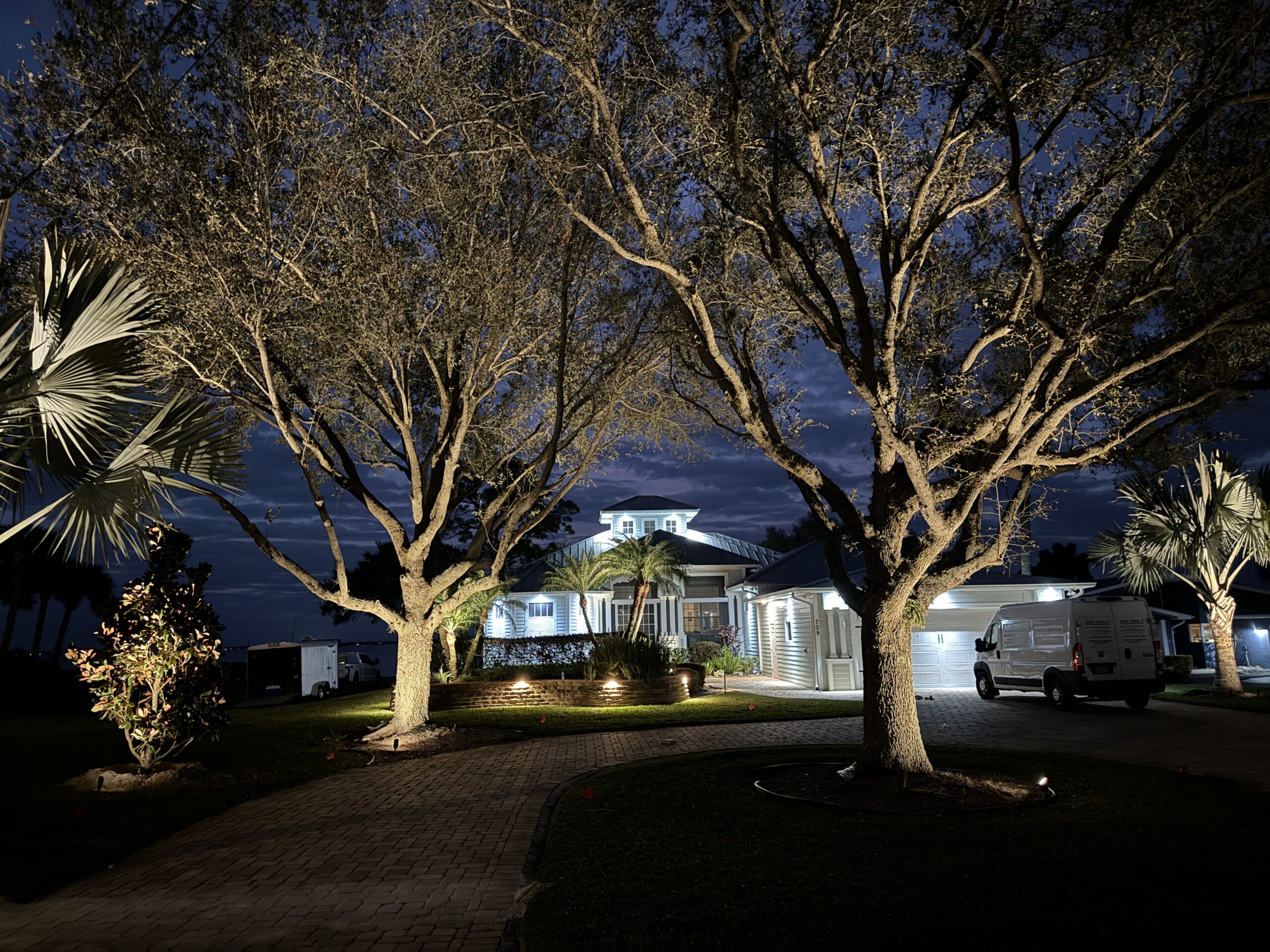 Large house at night illuminated by exterior lights, with two leafless trees in the foreground and a white van parked in the driveway.